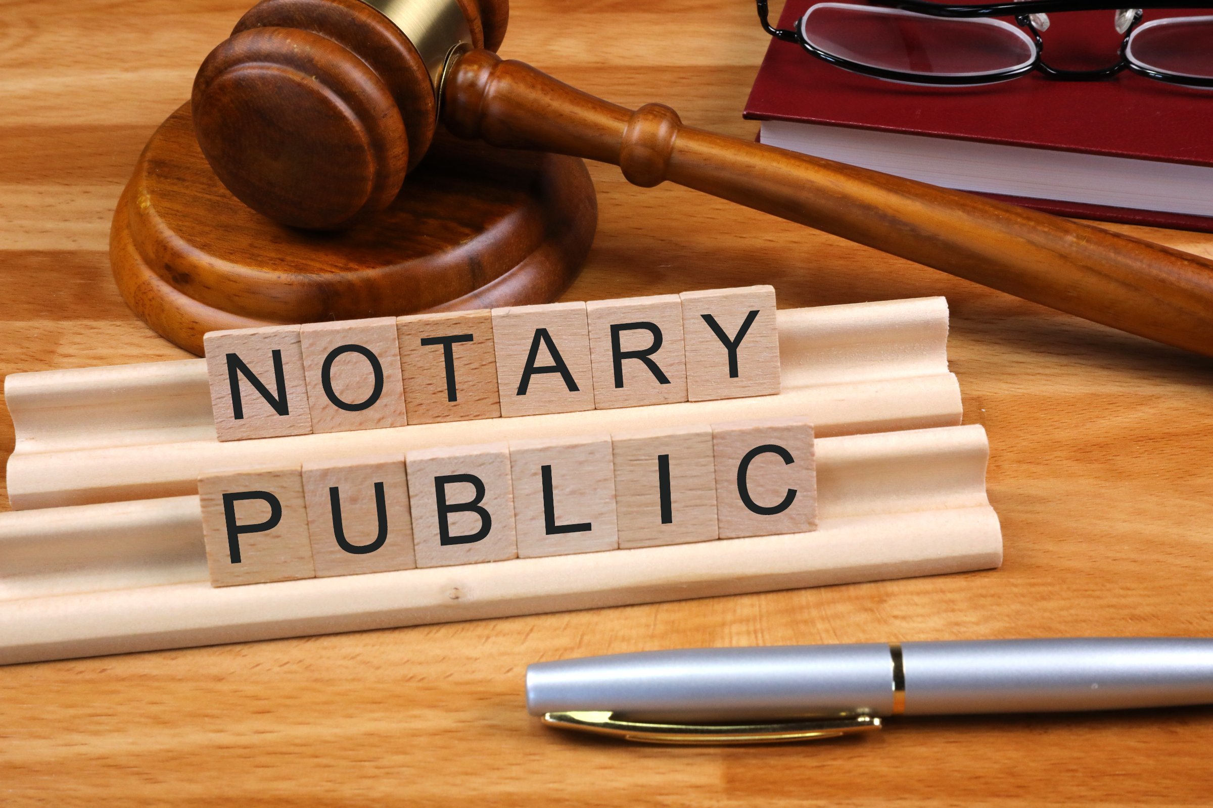 photo of the word notary public spelled out in individual scrabble tiles with a gavel behind and a silver pen in front.